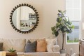 Beige living room with a round bulb border mirror above a sofa and a tall potted plant by a window.