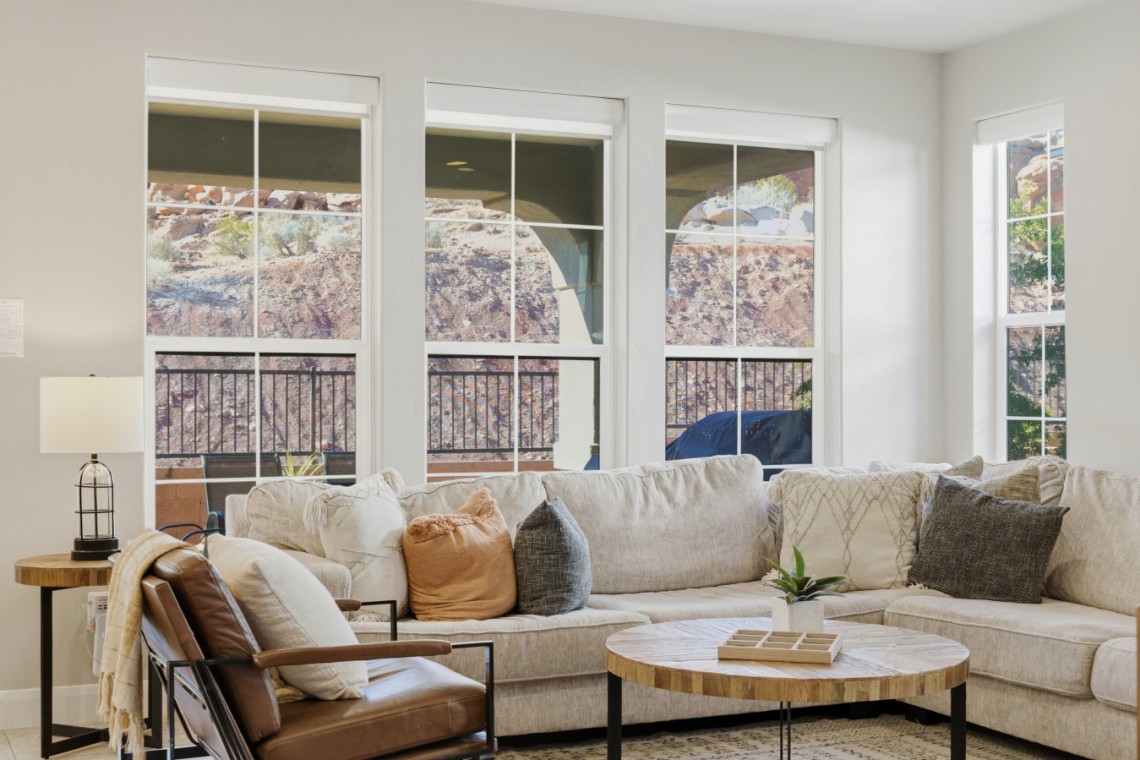 Bright living room with a beige sectional, multiple throw pillows, and a round wooden coffee table near large windows overlooking a rocky outdoor area.