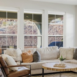 Bright living room with a beige sectional, multiple throw pillows, and a round wooden coffee table near large windows overlooking a rocky outdoor area.