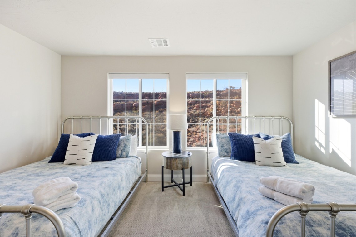 Twin bedroom with metal-frame beds, blue bedding, and a round nightstand with lamp between windows overlooking rocky hills outside.