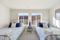 Twin bedroom with metal-frame beds, blue bedding, and a round nightstand with lamp between windows overlooking rocky hills outside.