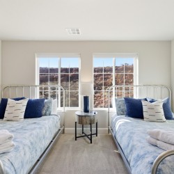 Twin bedroom with metal-frame beds, blue bedding, and a round nightstand with lamp between windows overlooking rocky hills outside.