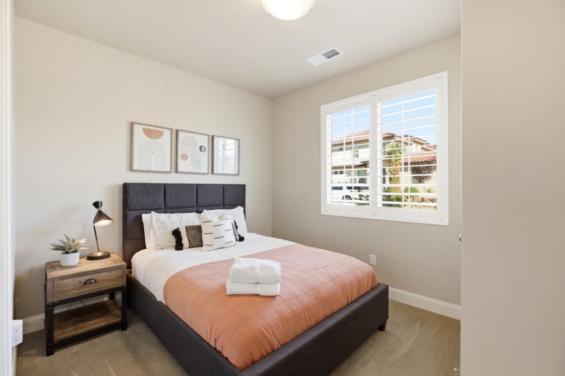 Cozy bedroom with a dark upholstered bed, peach quilt, white pillows, and a wooden nightstand with a lamp and plant by the window.