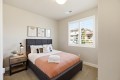 Cozy bedroom with a dark upholstered bed, peach quilt, white pillows, and a wooden nightstand with a lamp and plant by the window.