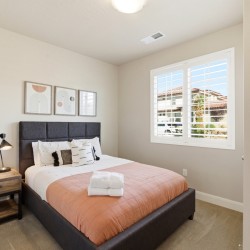 Cozy bedroom with a dark upholstered bed, peach quilt, white pillows, and a wooden nightstand with a lamp and plant by the window.