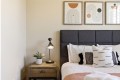 A bedside table with a plant and lamp sits beside a bed with a dark upholstered headboard and white bedding with decorative pillows; abstract art hangs above bed.