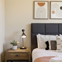 A bedside table with a plant and lamp sits beside a bed with a dark upholstered headboard and white bedding with decorative pillows; abstract art hangs above bed.