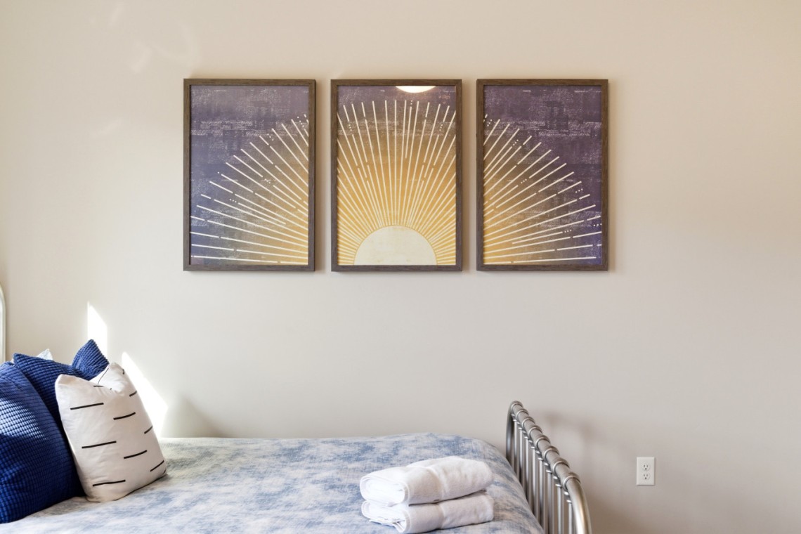Three-panel sunburst artwork hung above a bed with blue pillows and folded towels on the blanket.