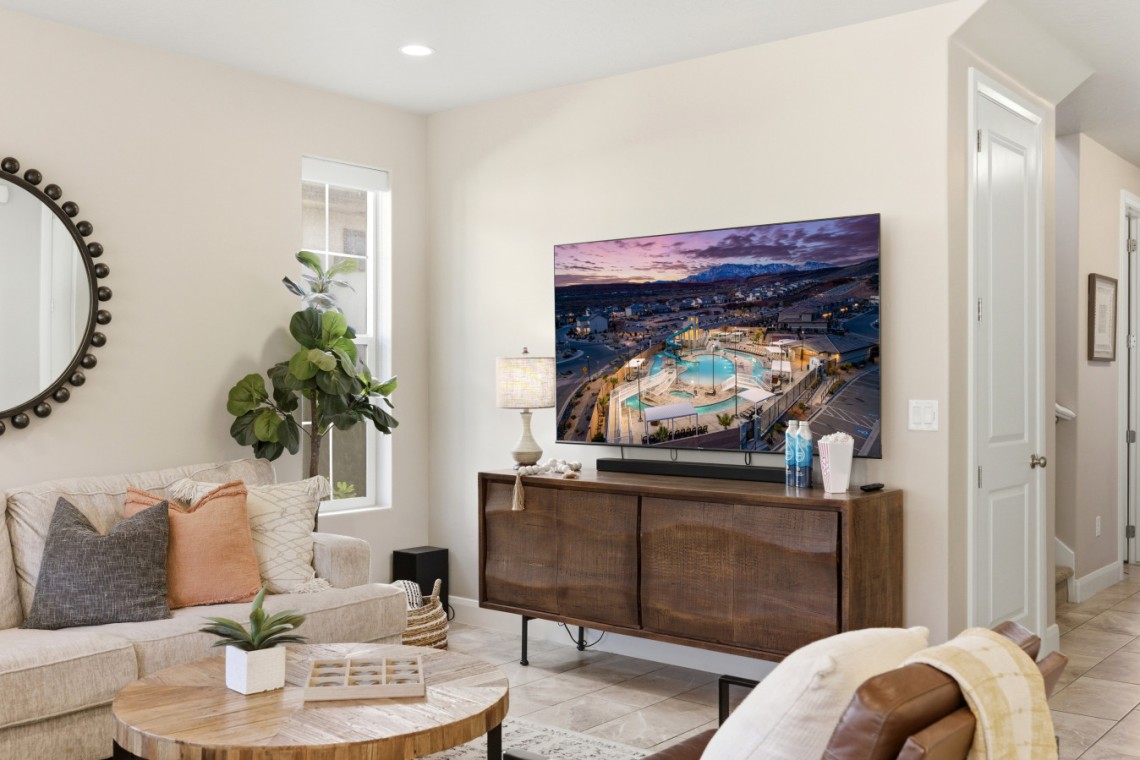 Modern living room with a wall-mounted flat-screen TV above a wooden media console, beige sofa with throw pillows, and a round-framed mirror on the wall.