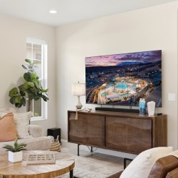 Modern living room with a wall-mounted flat-screen TV above a wooden media console, beige sofa with throw pillows, and a round-framed mirror on the wall.