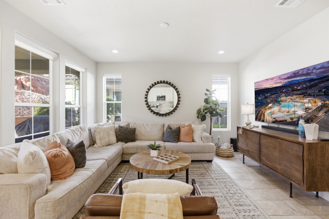 Bright living room with a beige L-shaped sofa, round wooden coffee table, and a large wall-mounted TV on a wooden console.