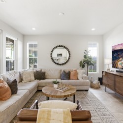 Bright living room with a beige L-shaped sofa, round wooden coffee table, and a large wall-mounted TV on a wooden console.