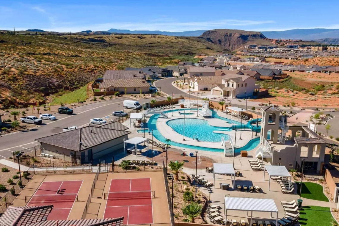 Aerial view of a desert resort-like community with a large central swimming pool, water slide, lounge chairs, and surrounding houses.