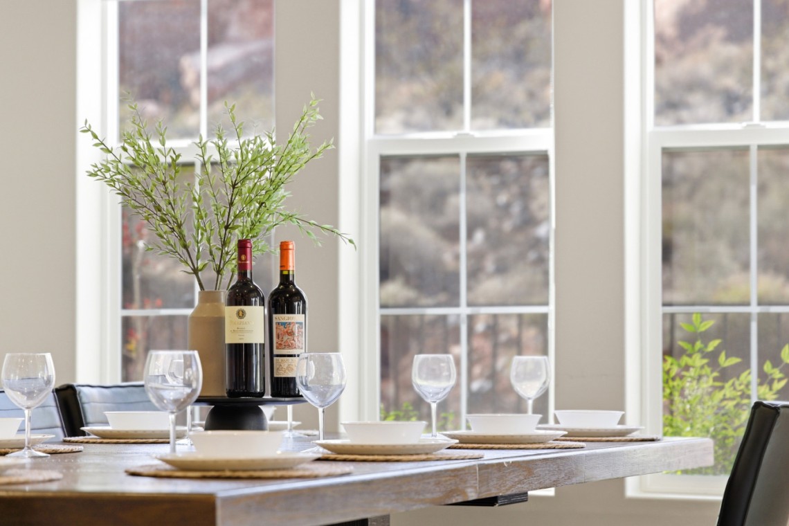 Elegant dining table set with two wine bottles as a centerpiece, surrounded by wine glasses and white dishes, near large windows with greenery outside.