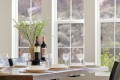 Elegant dining table set with two wine bottles as a centerpiece, surrounded by wine glasses and white dishes, near large windows with greenery outside.