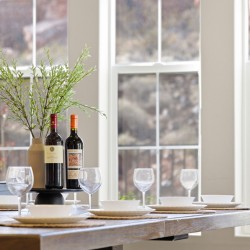 Elegant dining table set with two wine bottles as a centerpiece, surrounded by wine glasses and white dishes, near large windows with greenery outside.