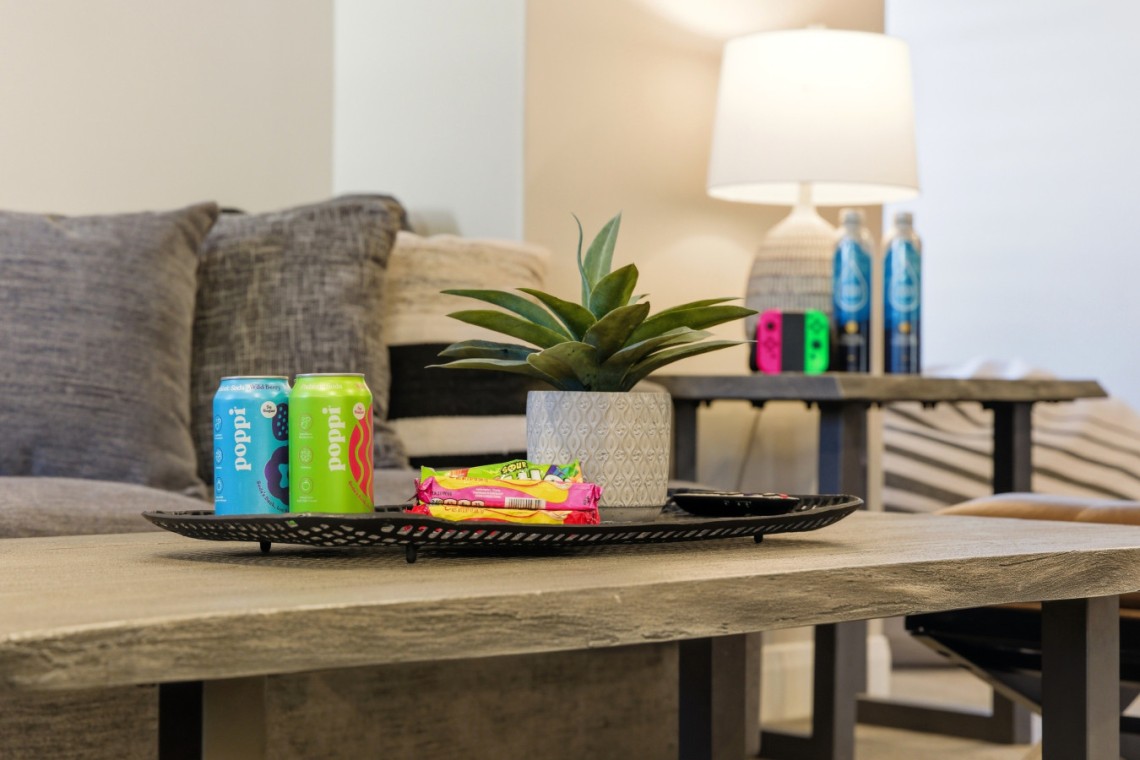 Living room coffee table with a metal tray holding two Poppi cans, snack bars, a potted plant, and a TV remote; sofa and lamp in the background.