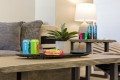 Living room coffee table with a metal tray holding two Poppi cans, snack bars, a potted plant, and a TV remote; sofa and lamp in the background.