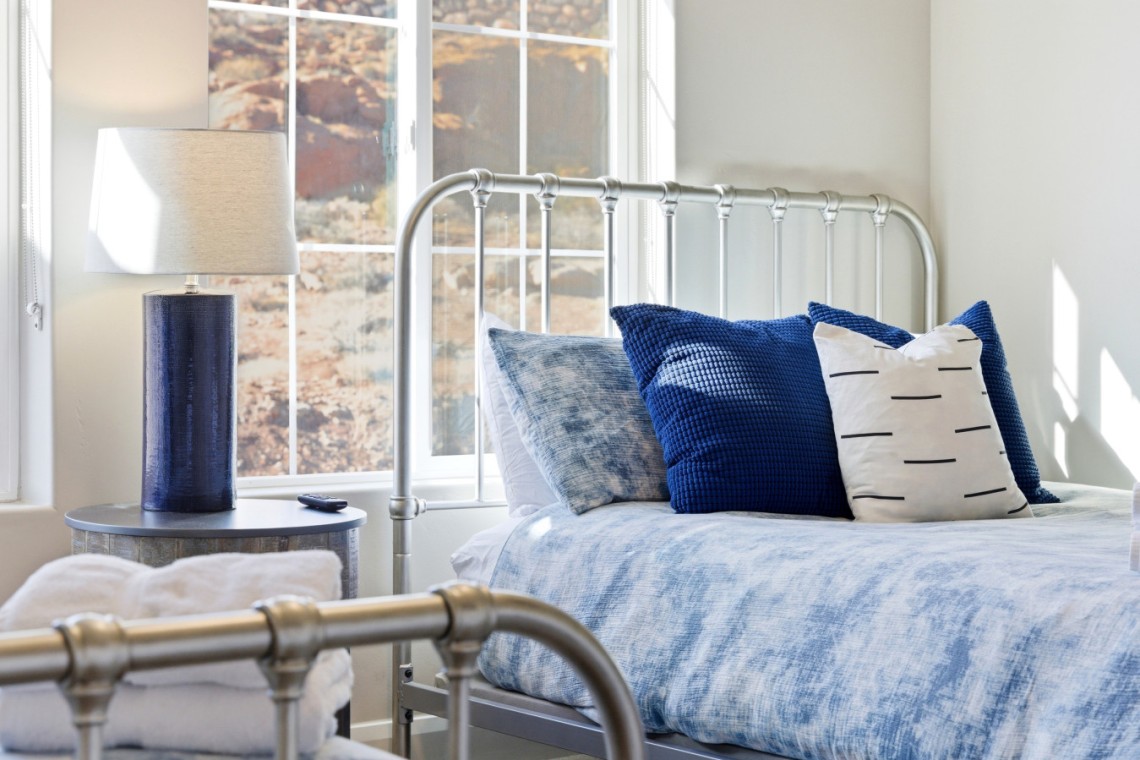 Twin metal bed with blue bedding and matching pillows beside a round blue-lamp table by a sunlit window.