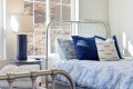 Twin metal bed with blue bedding and matching pillows beside a round blue-lamp table by a sunlit window.