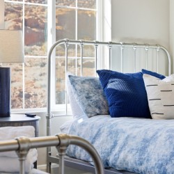 Twin metal bed with blue bedding and matching pillows beside a round blue-lamp table by a sunlit window.