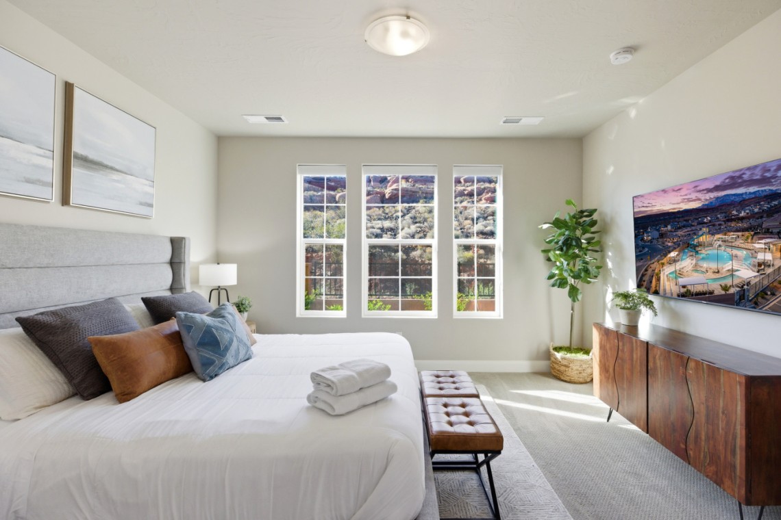 Bright modern bedroom with a large bed, gray headboard, and decorative pillows facing three tall windows letting in natural light. A brown bench sits at the foot of the bed.