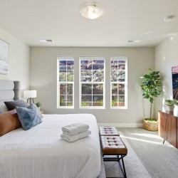 Bright modern bedroom with a large bed, gray headboard, and decorative pillows facing three tall windows letting in natural light. A brown bench sits at the foot of the bed.