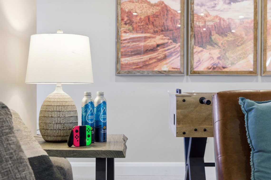 Living room scene with a ceramic lamp on a side table, two water bottles, and a Nintendo Switch with red and green Joy-Cons; a foosball table in the background.