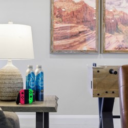 Living room scene with a ceramic lamp on a side table, two water bottles, and a Nintendo Switch with red and green Joy-Cons; a foosball table in the background.