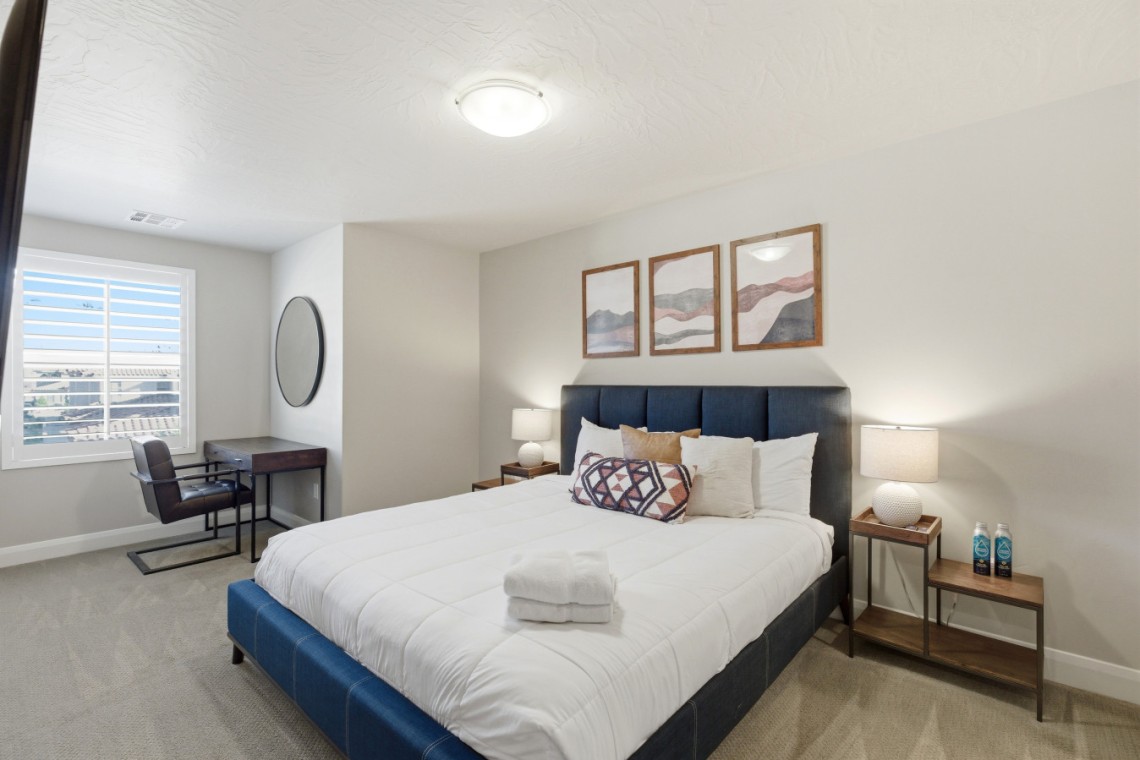 Hotel bedroom with a blue upholstered bed, white quilt, and two wood nightstands with lamps, plus three framed abstract landscape prints above the headboard.