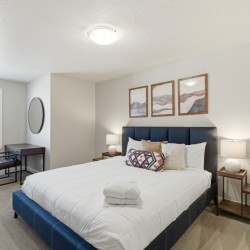 Hotel bedroom with a blue upholstered bed, white quilt, and two wood nightstands with lamps, plus three framed abstract landscape prints above the headboard.