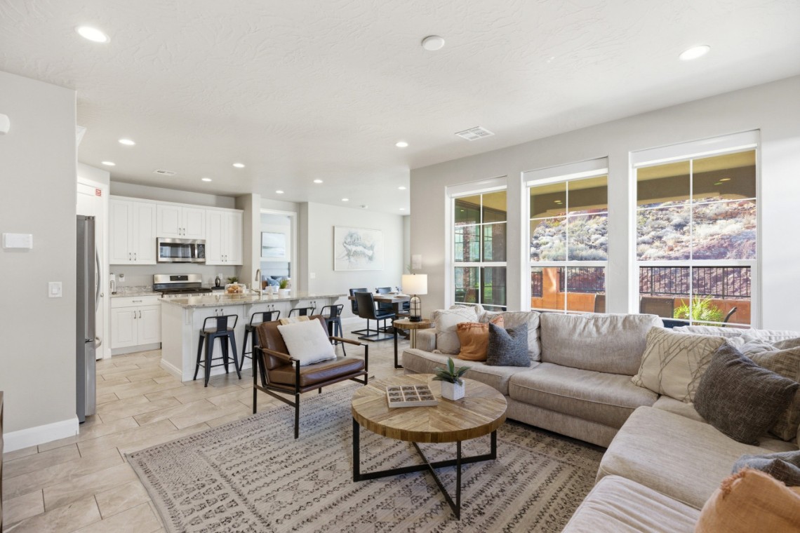 Open-concept living room and kitchen with a beige sectional, round wooden coffee table, and large windows overlooking a patio.