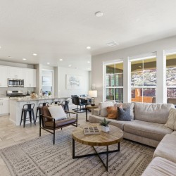 Open-concept living room and kitchen with a beige sectional, round wooden coffee table, and large windows overlooking a patio.