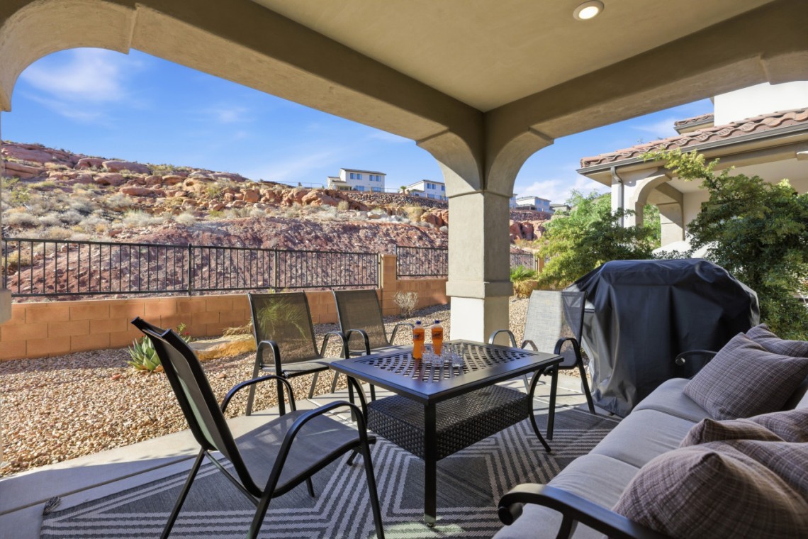 Covered patio with a metal dining table and six chairs, a cushioned couch, and a covered grill, overlooking a rocky desert yard.
