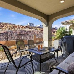 Covered patio with a metal dining table and six chairs, a cushioned couch, and a covered grill, overlooking a rocky desert yard.