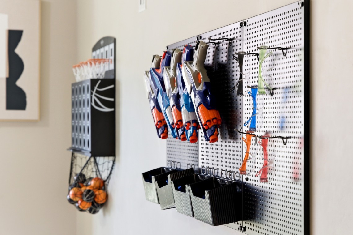 Pegboard wall with blue and orange Nerf-style toy guns hung on hooks, with black bins underneath; a wire basket of orange balls hangs to the left.