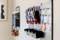 Pegboard wall with blue and orange Nerf-style toy guns hung on hooks, with black bins underneath; a wire basket of orange balls hangs to the left.