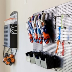 Pegboard wall with blue and orange Nerf-style toy guns hung on hooks, with black bins underneath; a wire basket of orange balls hangs to the left.