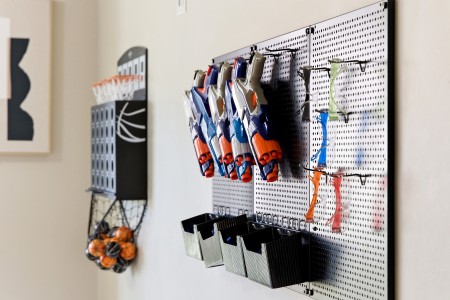 Pegboard wall with blue and orange Nerf-style toy guns hung on hooks, with black bins underneath; a wire basket of orange balls hangs to the left.