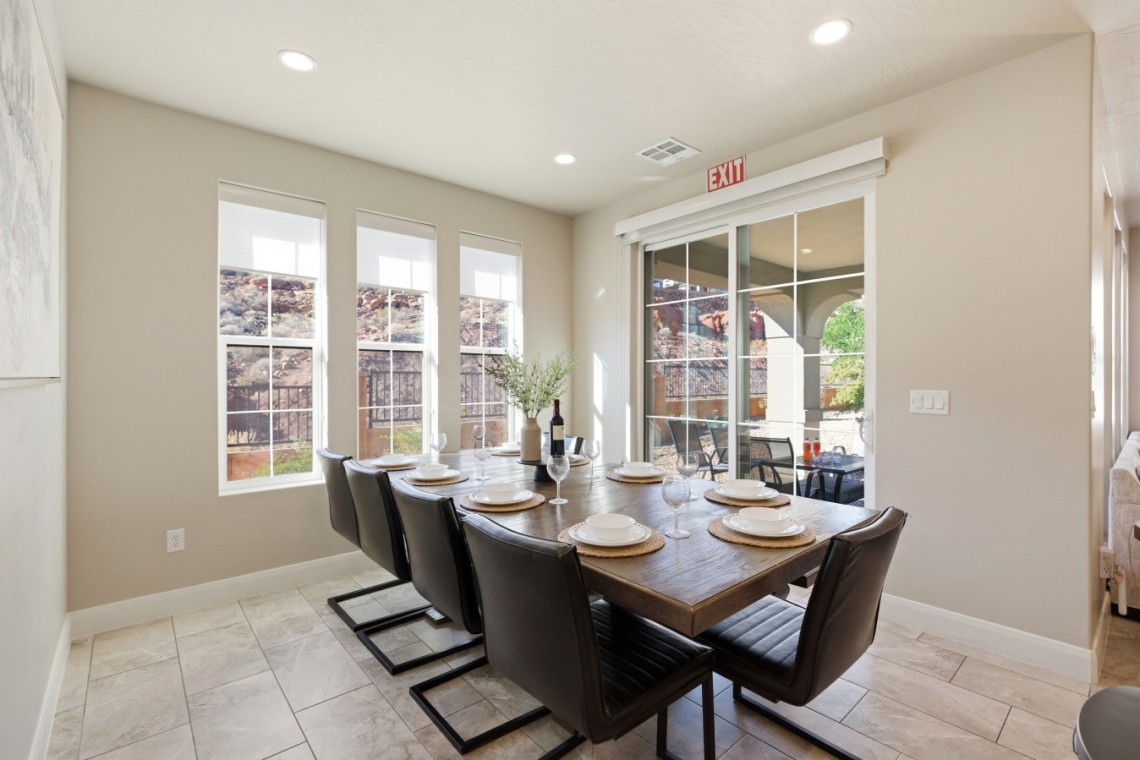 Dining room with a long wooden table set for six, black leather chairs, and a wine bottle centerpiece by a row of windows.