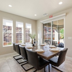 Dining room with a long wooden table set for six, black leather chairs, and a wine bottle centerpiece by a row of windows.