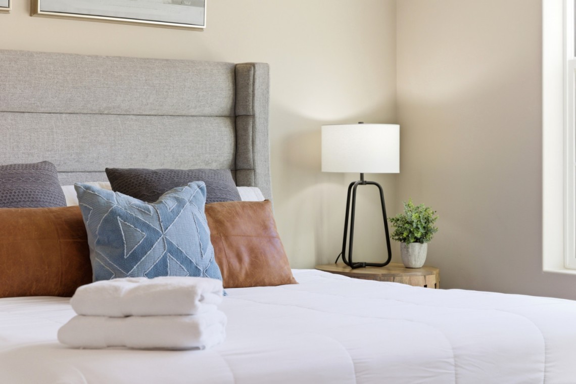 Bedroom with a padded gray headboard, decorative pillows, and a stack of white towels on a white comforter.