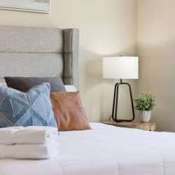Bedroom with a padded gray headboard, decorative pillows, and a stack of white towels on a white comforter.