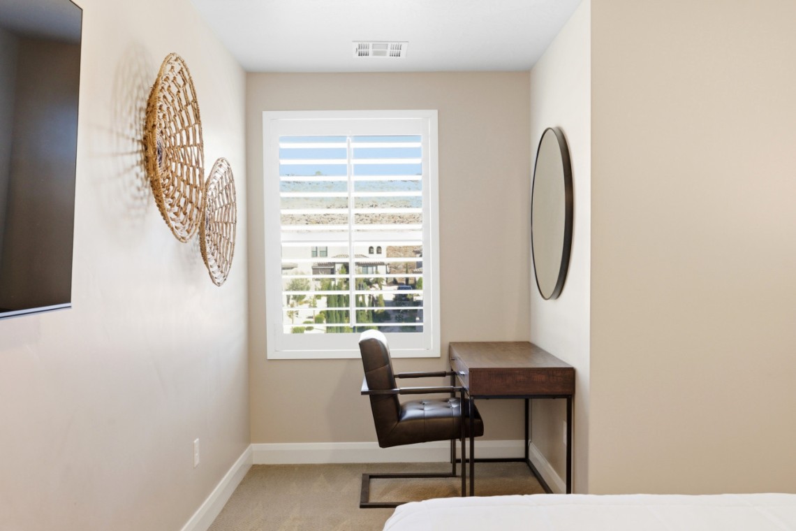 A small home office with a wooden desk and brown leather chair by a window with white blinds; woven circular wall art on the left and a round mirror on the right