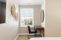 A small home office with a wooden desk and brown leather chair by a window with white blinds; woven circular wall art on the left and a round mirror on the right