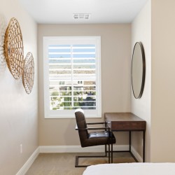 A small home office with a wooden desk and brown leather chair by a window with white blinds; woven circular wall art on the left and a round mirror on the right