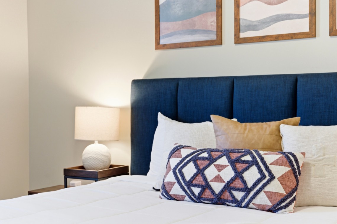 Bedroom scene with a navy-blue upholstered headboard, white bedding, and a geometric-patterned throw pillow in front of several neutral pillows; a bedside table and lamp on the left.