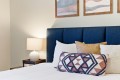 Bedroom scene with a navy-blue upholstered headboard, white bedding, and a geometric-patterned throw pillow in front of several neutral pillows; a bedside table and lamp on the left.