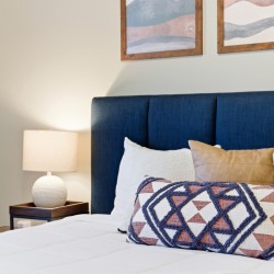 Bedroom scene with a navy-blue upholstered headboard, white bedding, and a geometric-patterned throw pillow in front of several neutral pillows; a bedside table and lamp on the left.