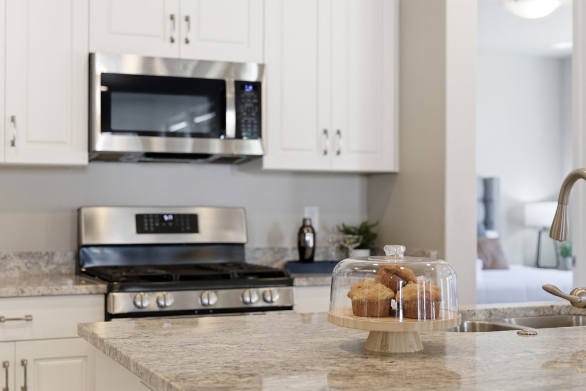 Muffins under a glass cloche on a wooden pedestal atop a granite kitchen island, with stainless steel appliances in the background.